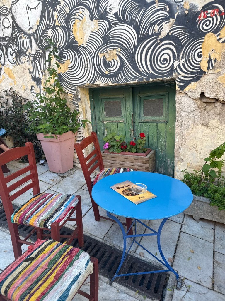 A blue table with a magazine and an empty ashtray, surrounded by colorful chairs, in front of a textured wall featuring swirling black and white art and green doors.