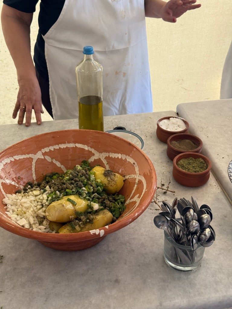 A mixed bowl of diced potatoes, chopped green onions, and capers with herbs and spices on a marble countertop, accompanied by a bottle of olive oil and containers of salt and spices.