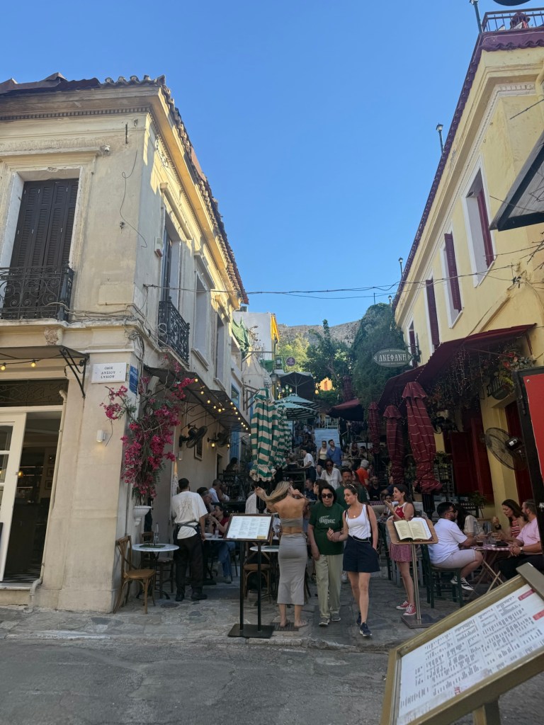 A bustling street scene in a vibrant, narrow alleyway lined with restaurants and cafes, filled with people dining and socializing under a clear blue sky.