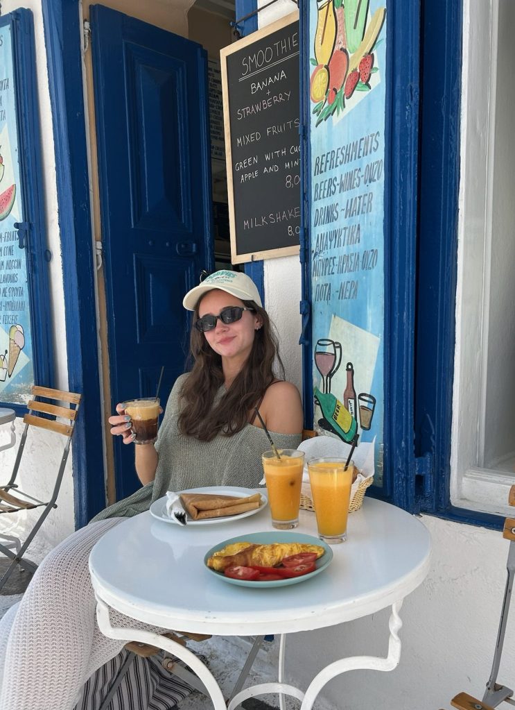 A woman sitting at a white table outside a café, holding a drink, with two fruit juices and a plate of food in front of her, colorful café signage in the background.