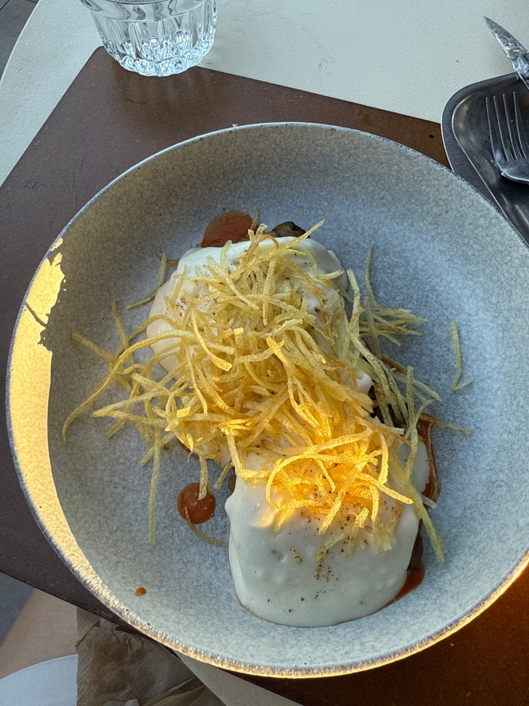 A plate of food featuring two items covered in a creamy white sauce, topped with golden crispy potato strips, with a small serving of sauce on the side. A glass of water is visible in the background.