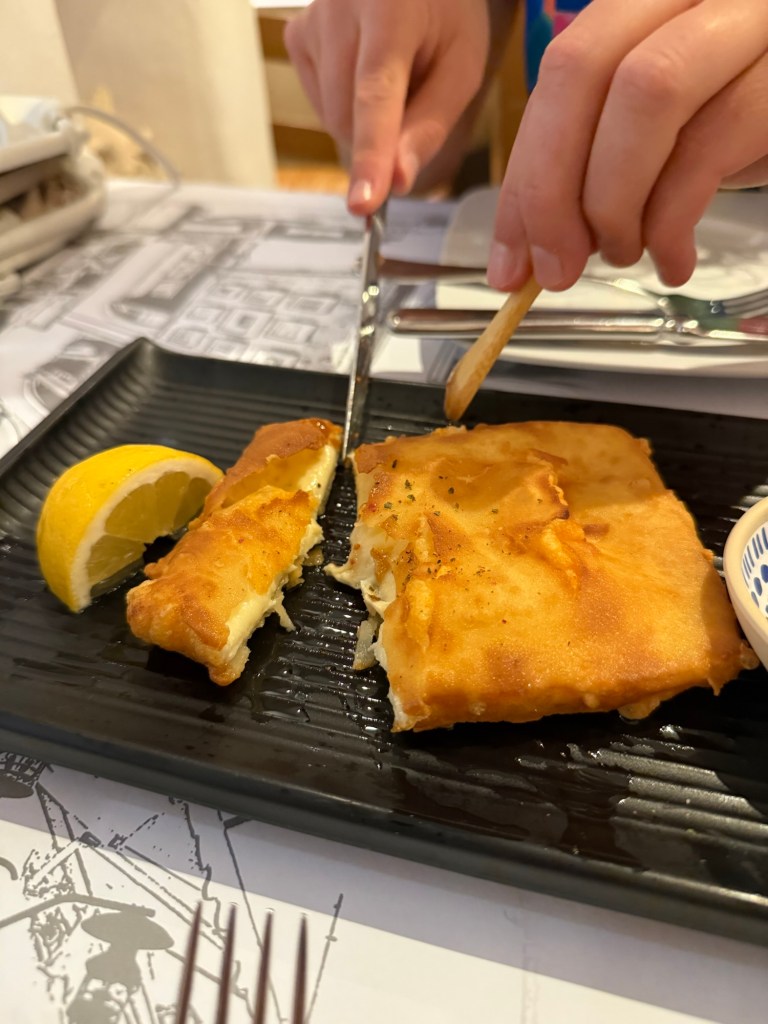 A close-up of a person using a knife and fork to cut a golden-brown fried dish on a black plate, accompanied by a slice of lemon.