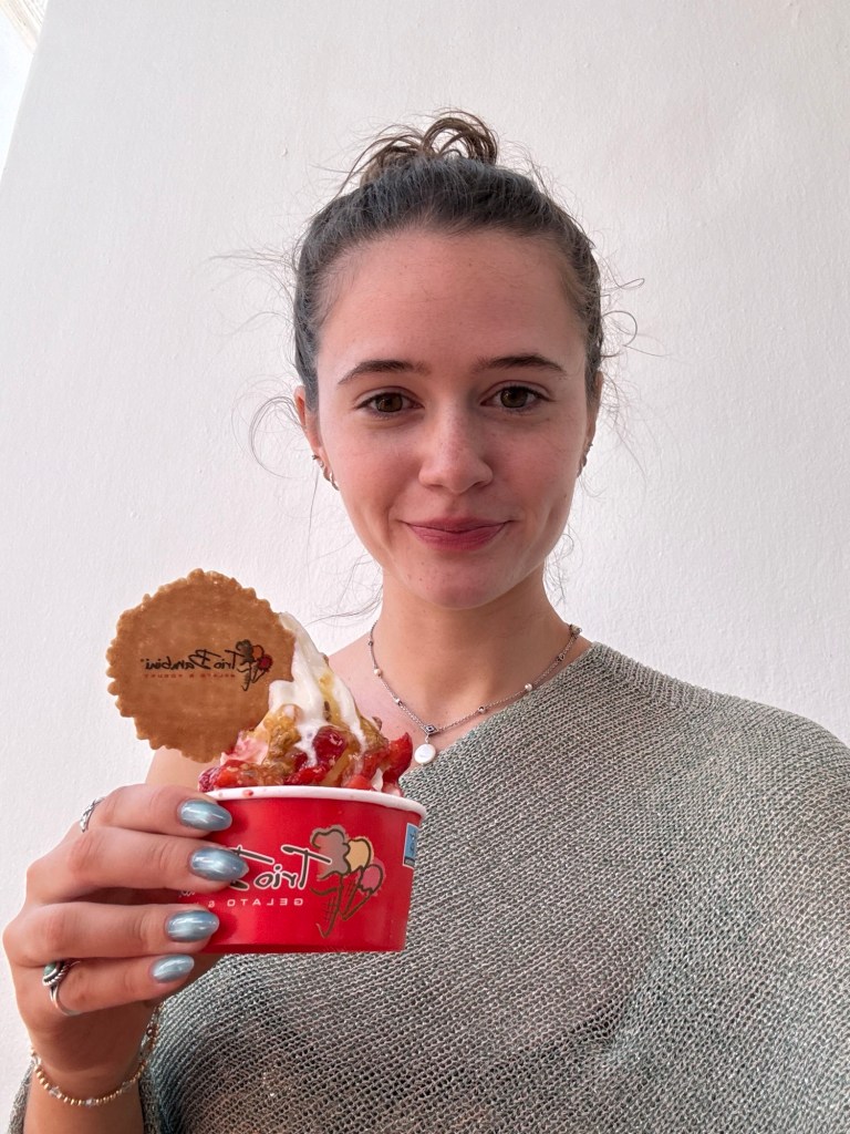 A young woman holding a cup of gelato topped with whipped cream and a decorative cookie, smiling at the camera.