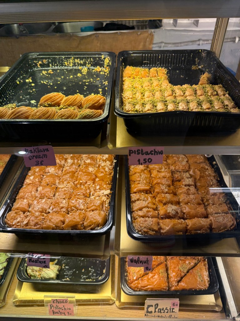 An assortment of baklava varieties displayed in a glass case, including pistachio, walnut, and cream baklava, with price tags visible.