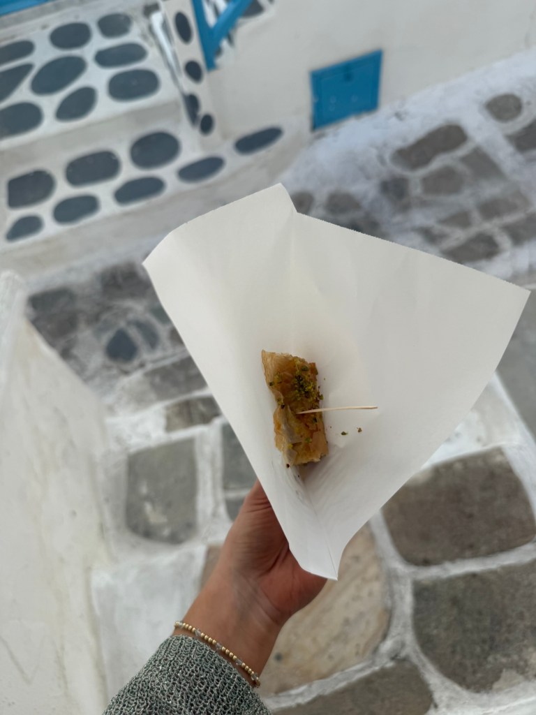 A close-up view of a hand holding a piece of baklava on a wooden toothpick, wrapped in white paper, with a blurred background of stone pavement and a blue and white building.