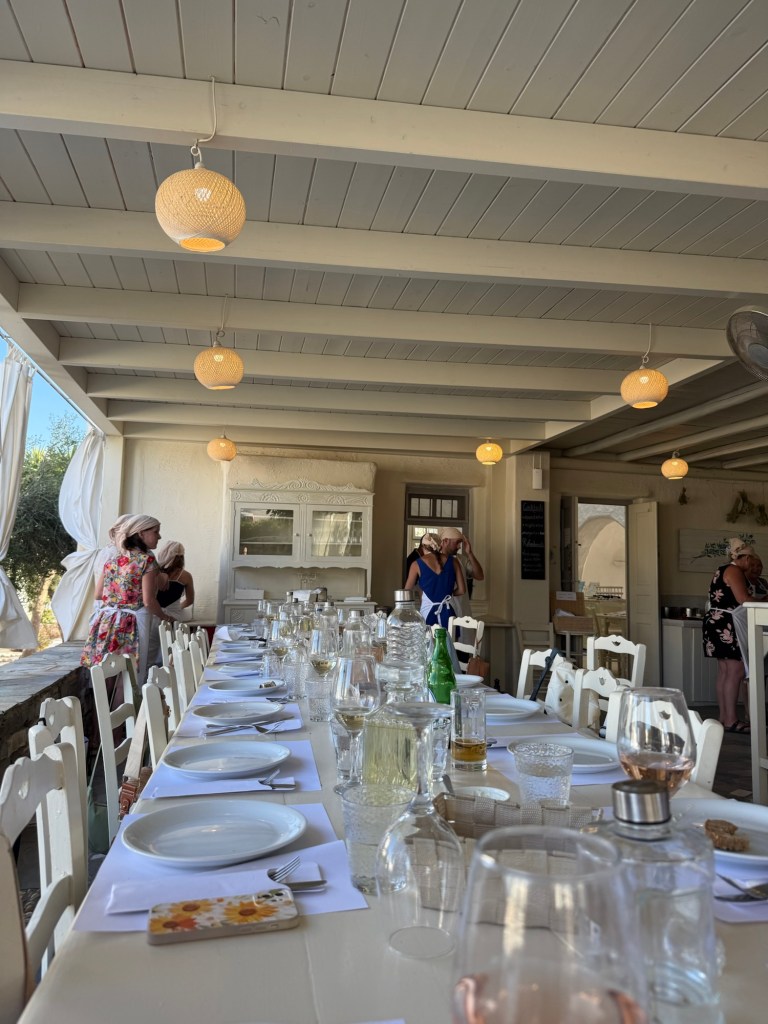 A long dining table set for a meal, featuring white plates and glassware, with decorative light fixtures hanging overhead. In the background, several people are engaged in conversation.