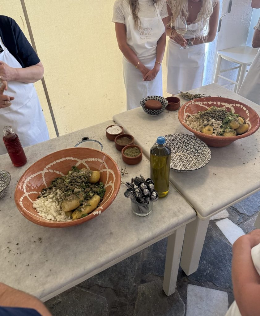 A cooking setup featuring a large terracotta bowl with potatoes, greens, and various seasonings, surrounded by smaller bowls of spices and an olive oil bottle on a marble table, with participants in white aprons in the background.
