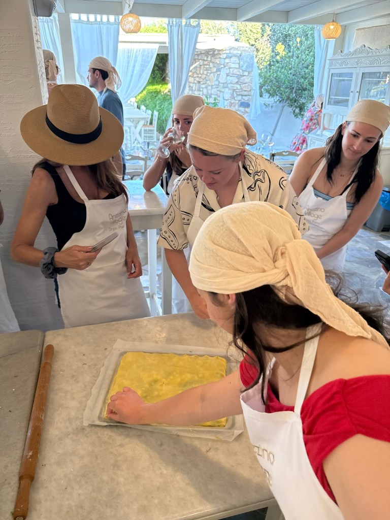 A group of people wearing aprons and head coverings are gathered around a table, engaged in a cooking activity. One person is spreading dough on parchment paper while others observe and socialize in a bright kitchen setting.
