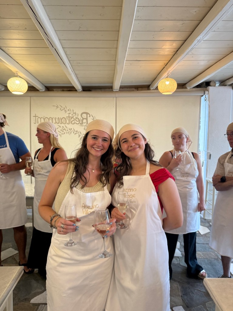 Two women posing together with glasses of rosé, wearing aprons and headbands, in a restaurant kitchen setting. Other participants are visible in the background.