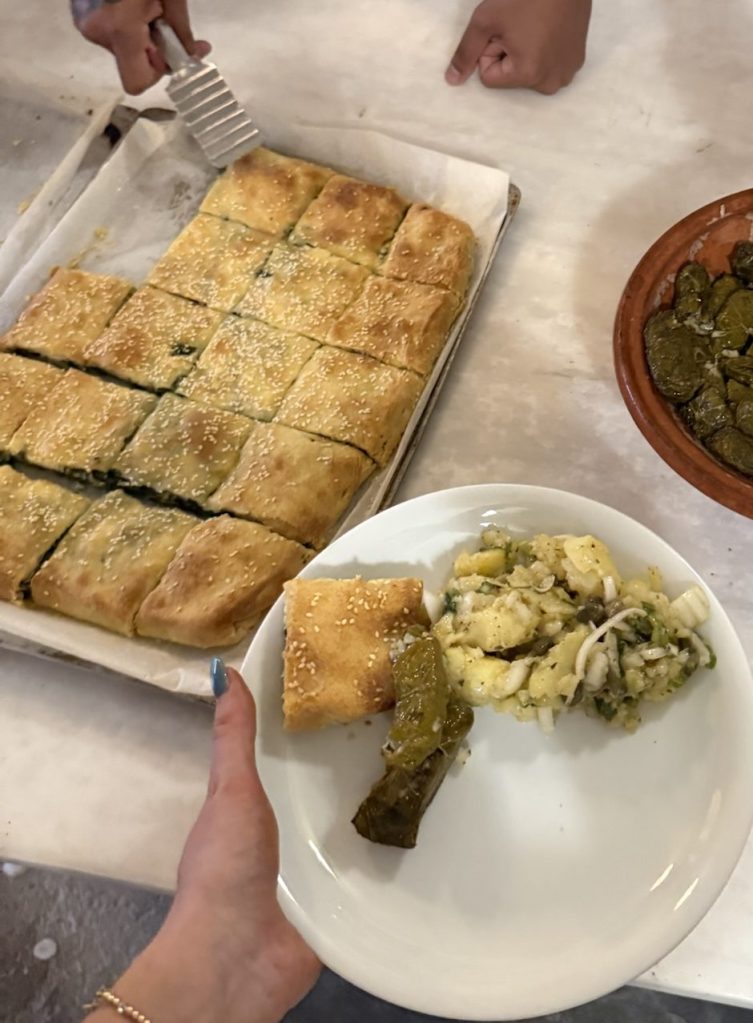 A person holding a plate of food containing a piece of pastry, a green pepper, and a potato salad, with a tray of sliced pastry in the background.