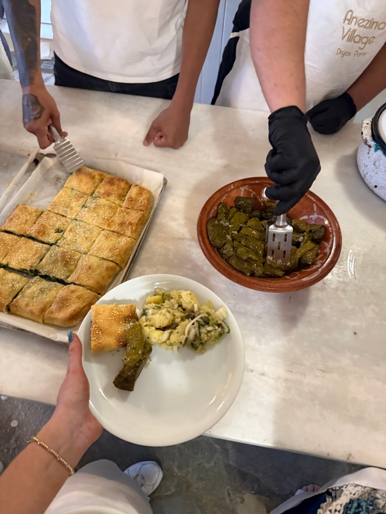 A person is serving traditional Mediterranean dishes, including a plate with stuffed grape leaves, a piece of baklava, and a vegetable medley, with additional baklava pieces in the background.