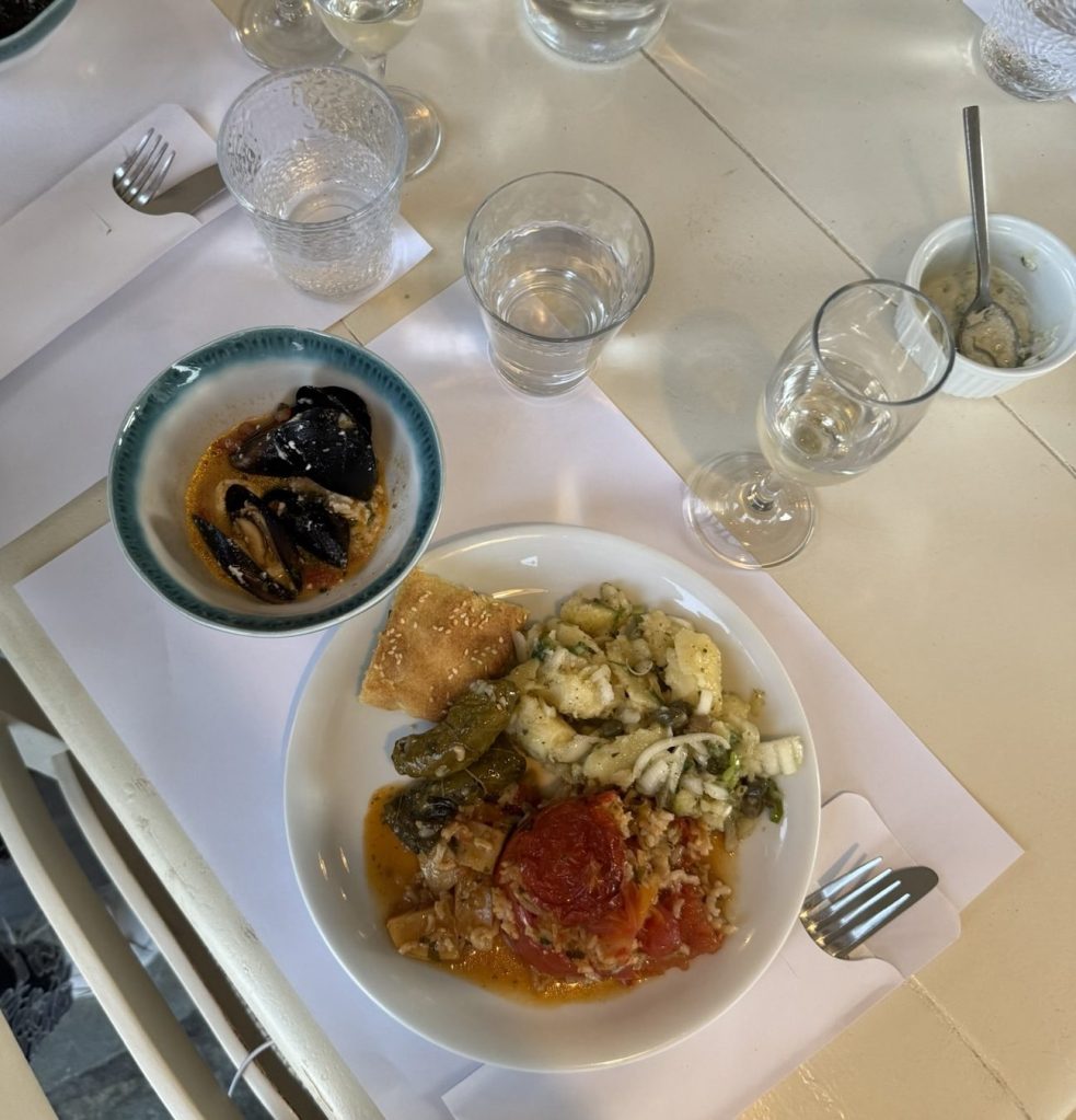 A plate of food featuring sliced tomatoes, green peppers, and a grain dish, accompanied by a piece of sesame bread. Nearby, a small bowl contains dark, glossy food items, and several glasses of water and wine are visible on the table.