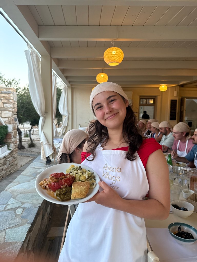A smiling young woman wearing an apron stands in a dining setting, holding a plate of traditional dishes, including stuffed vegetables and a side of rice. The background features other diners and decorative lights hanging from the ceiling.