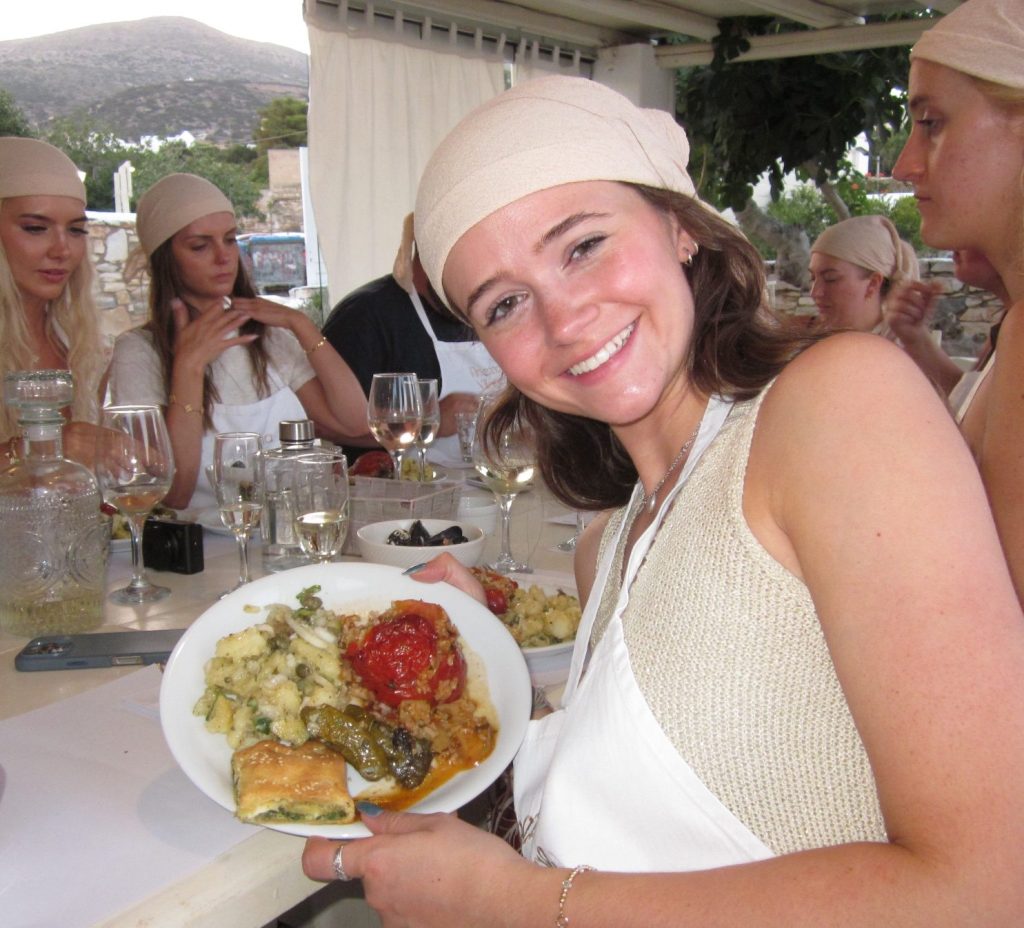 A young woman smiling and holding a plate of food, surrounded by other people at a dining table, with wine glasses and various dishes visible.